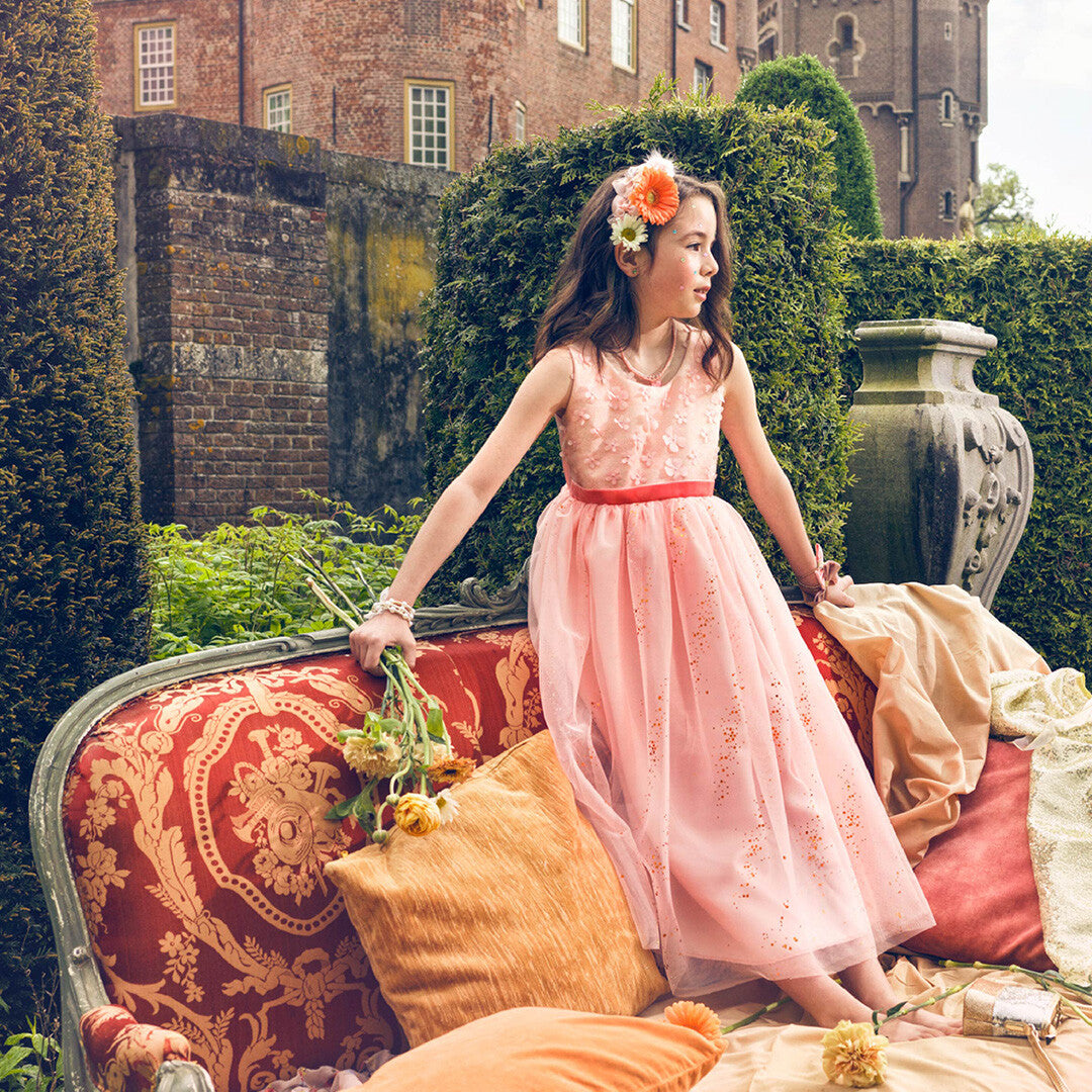 Een meisje in een roze kinderfeestjurk zit op een bank voor een kasteel.