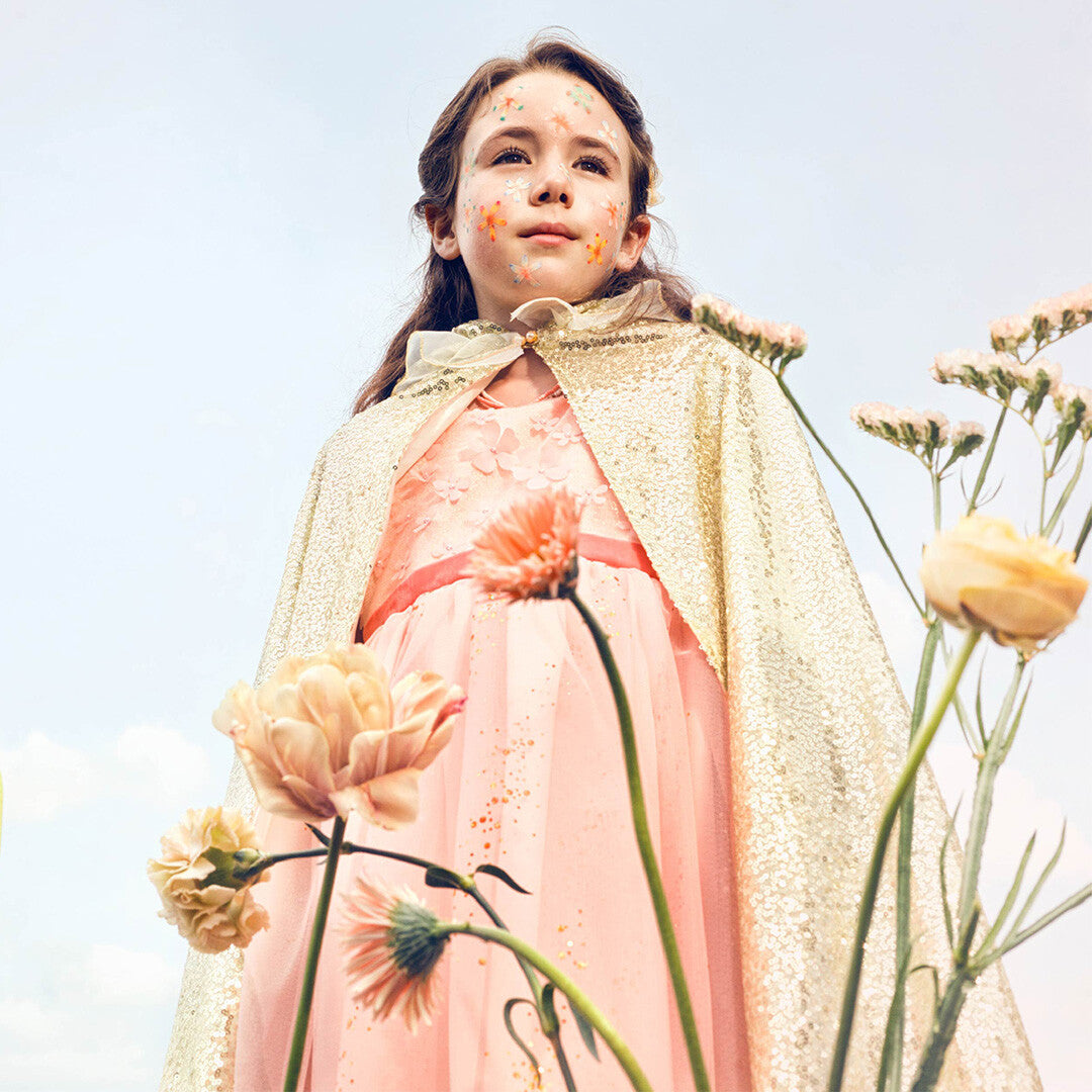 Een meisje in een roze kinderfeestjurk en gouden cape kijkt omhoog tussen bloemen.