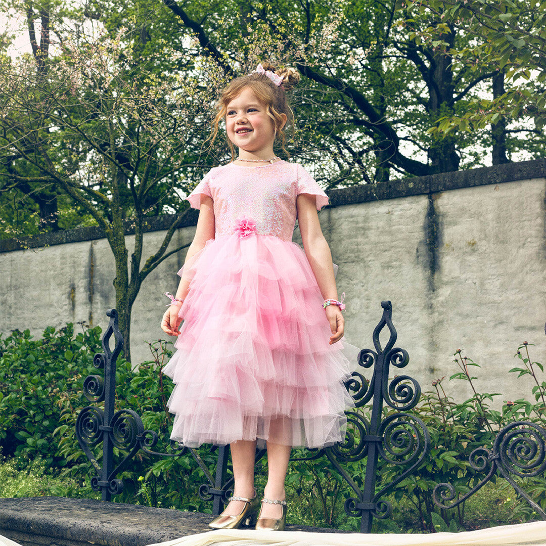 Een meisje draagt de roze prinsessen feestjurk en gouden schoenen, staand op een muurtje in een tuin.