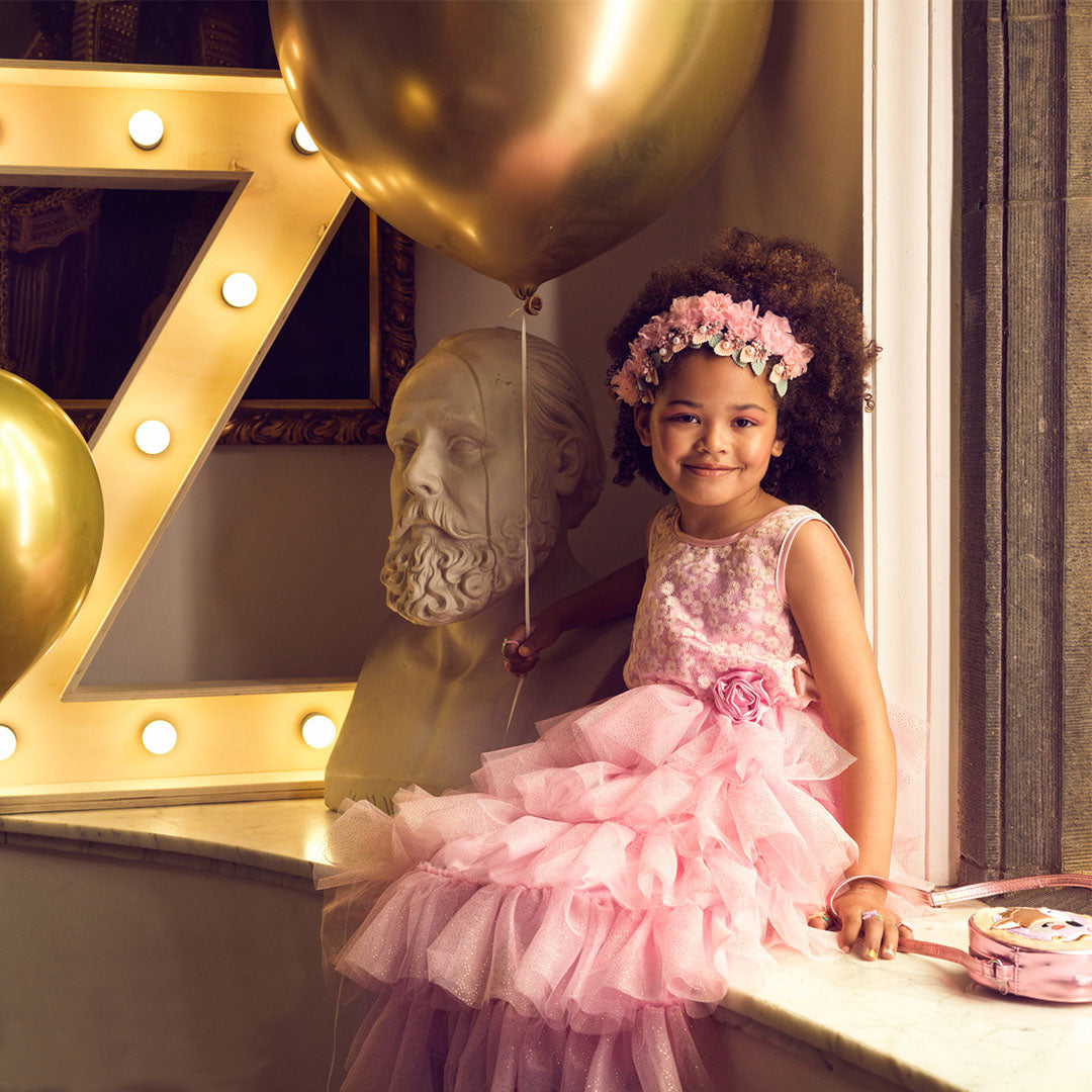 Een meisje met krullen en een bloemenkrans draagt de roze Souza for Kids prinsessenjurk en zit lachend op een vensterbank.