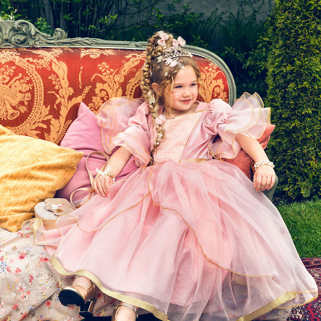 Meisje in een roze prinsessen kostuumjurk zit op een sierlijke bank in de tuin.
