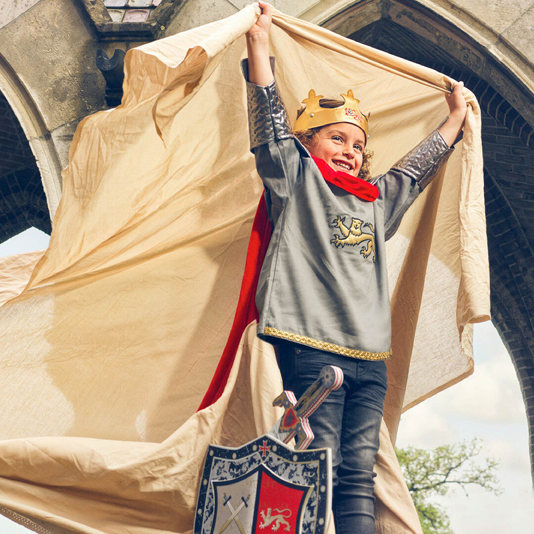 Een kind draagt het koning Arthur kinderkostuum met kroon, zwaard en schild, en houdt een beige doek omhoog voor een stenen boog.