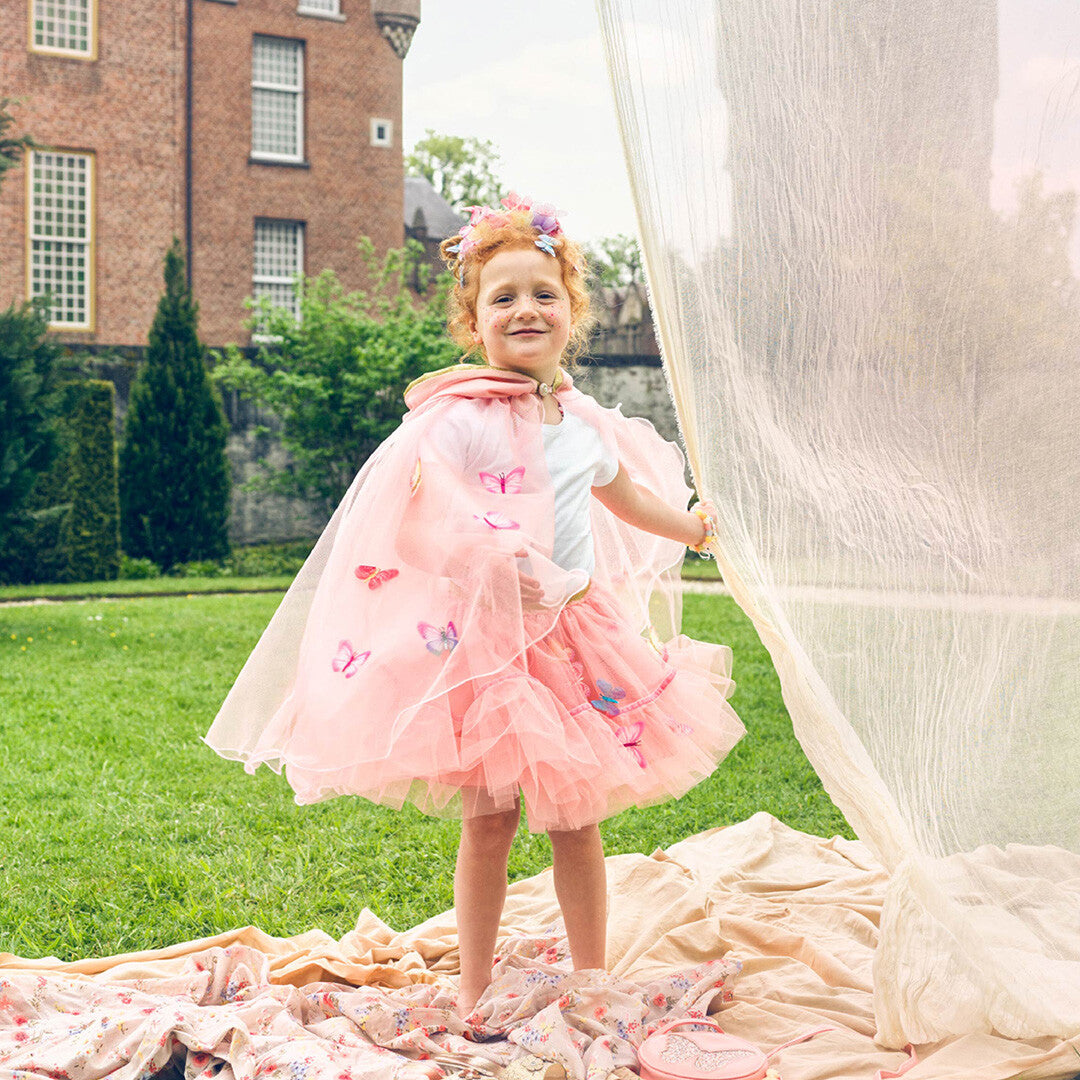 Een kind draagt de roze kinderrok met vlinders en een cape, staand op een deken in een tuin.