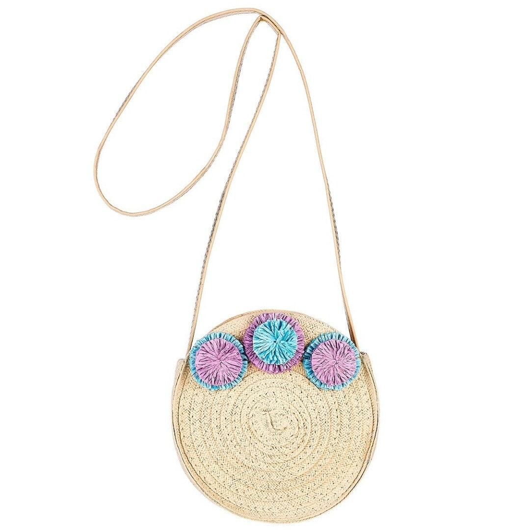 Souza for Kids ronde kinderschoudertas van beige vlechtwerk met paarse en blauwe pompons, schuin gefotografeerd.