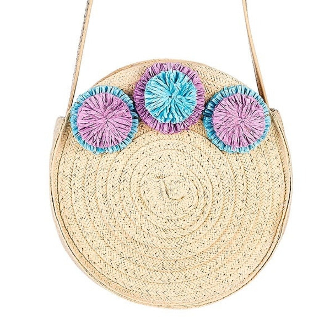 Souza for Kids ronde kinderschoudertas van beige vlechtwerk met drie paarse en blauwe pompons, van voren gefotografeerd.