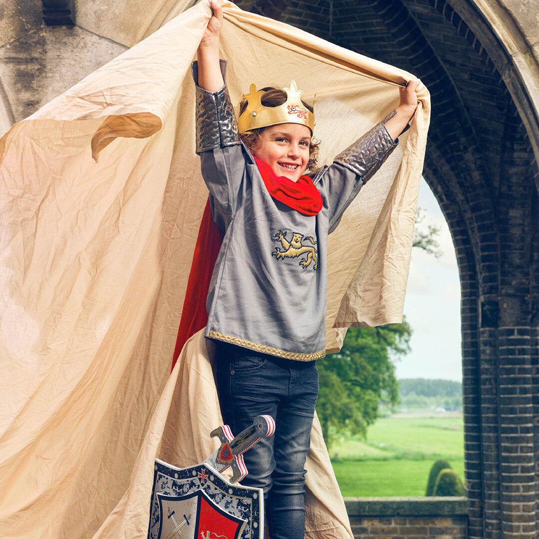 Een lachend kind verkleed als ridder of koning, met de gouden kroon, een cape, schild en zwaard, staand voor een kasteelmuur.