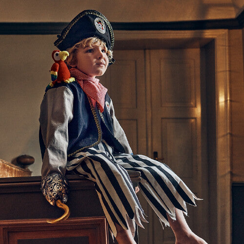 Een kind draagt het piratenkostuum met gestreepte broek, piratenhoed en haak, zittend op een meubel.