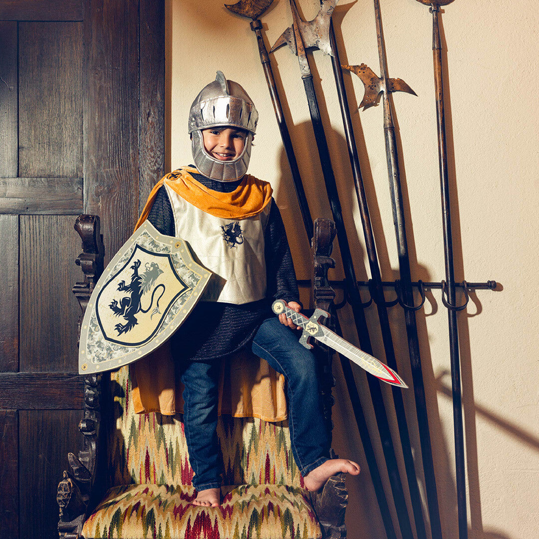 Een lachend kind in een ridderkostuum met zilveren helm, schild en zwaard, zittend op een stoel.