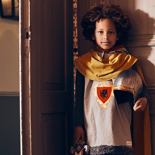 Een kind draagt het ridderkostuum met gele cape en houdt een helm en speelgoedzwaard vast.
