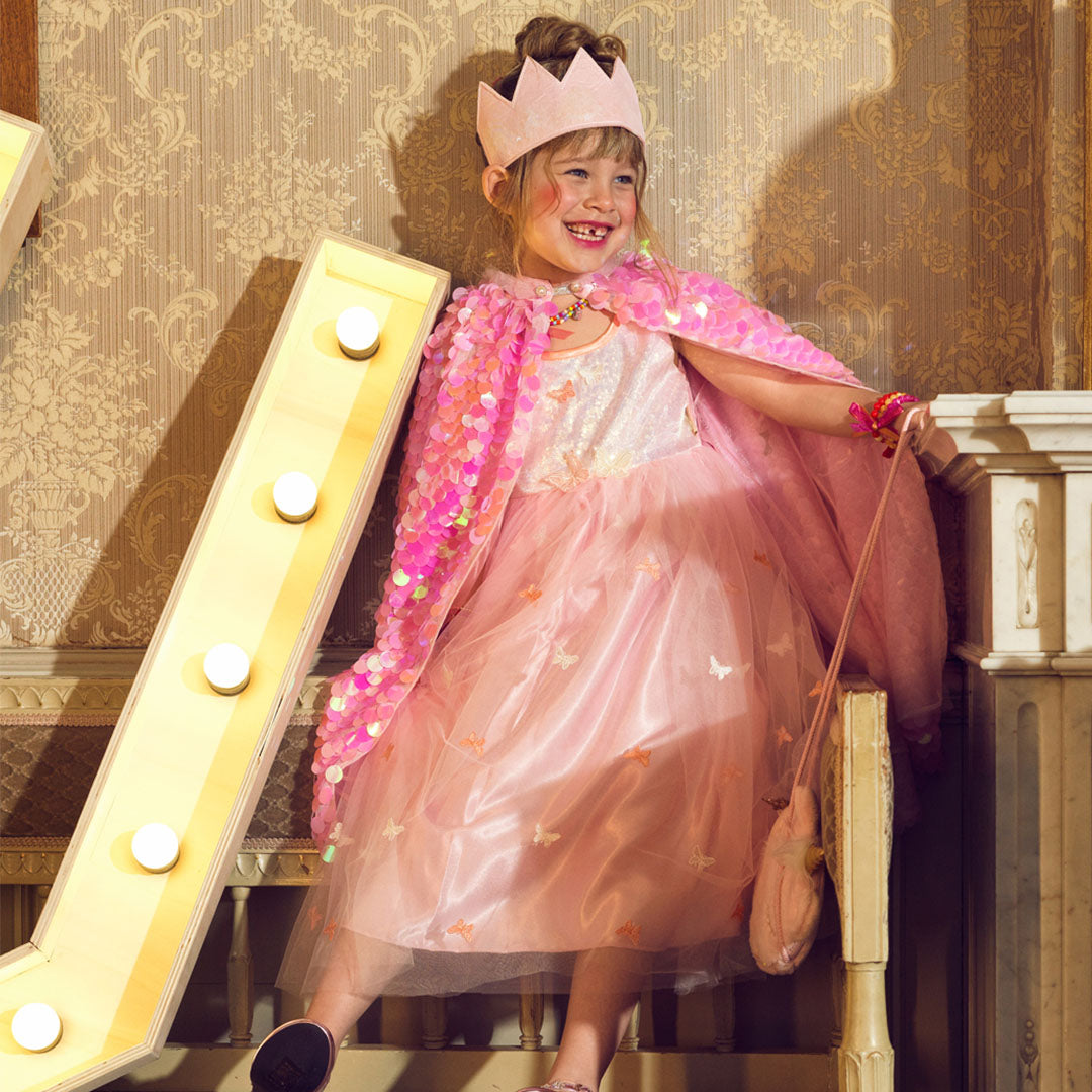 Een lachend meisje draagt de roze verkleedcape met schubben en een kroon, staand op een trap.