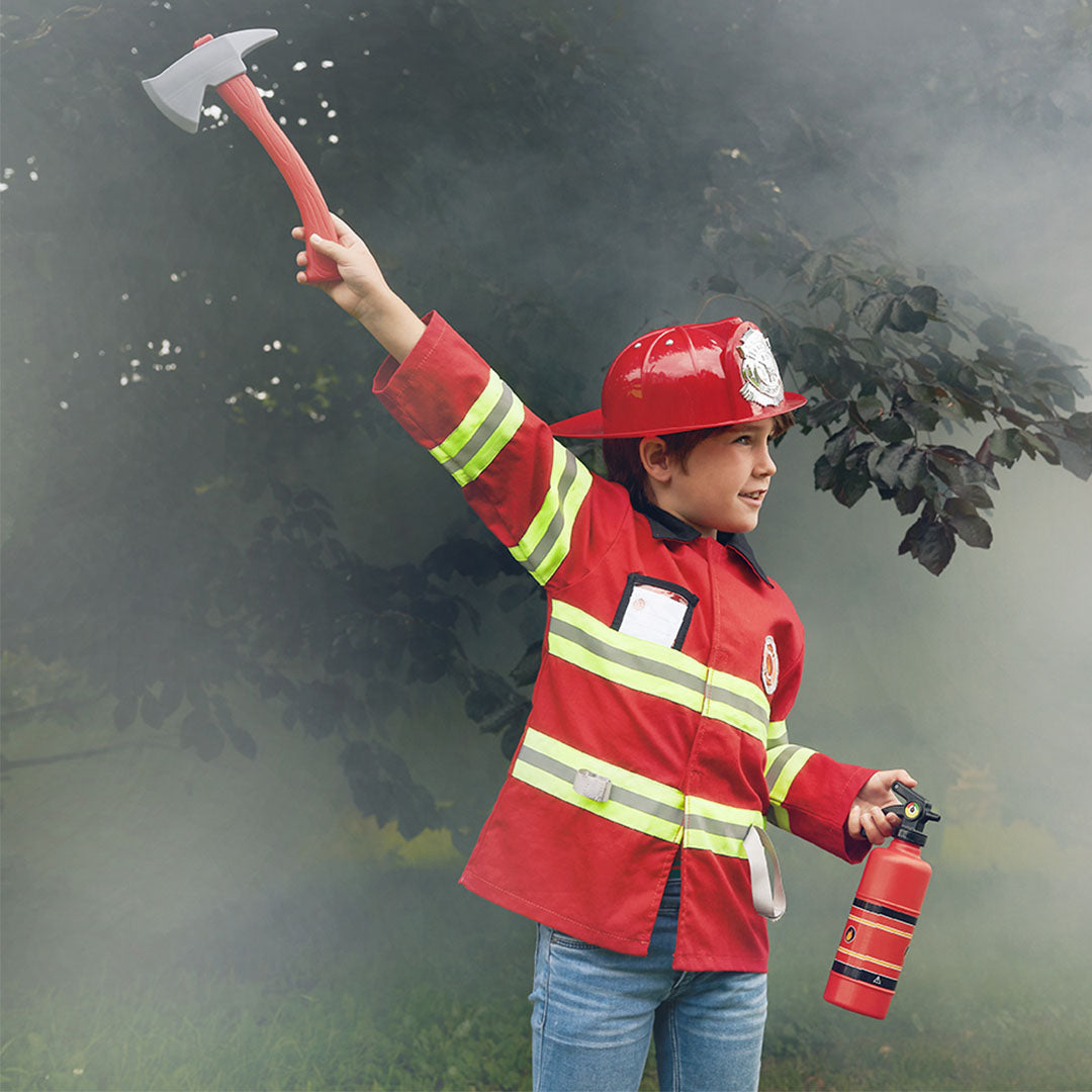 Een jongen in brandweer verkleedkleding met rode jas en helm houdt een speelgoedbijl en brandblusser omhoog.
