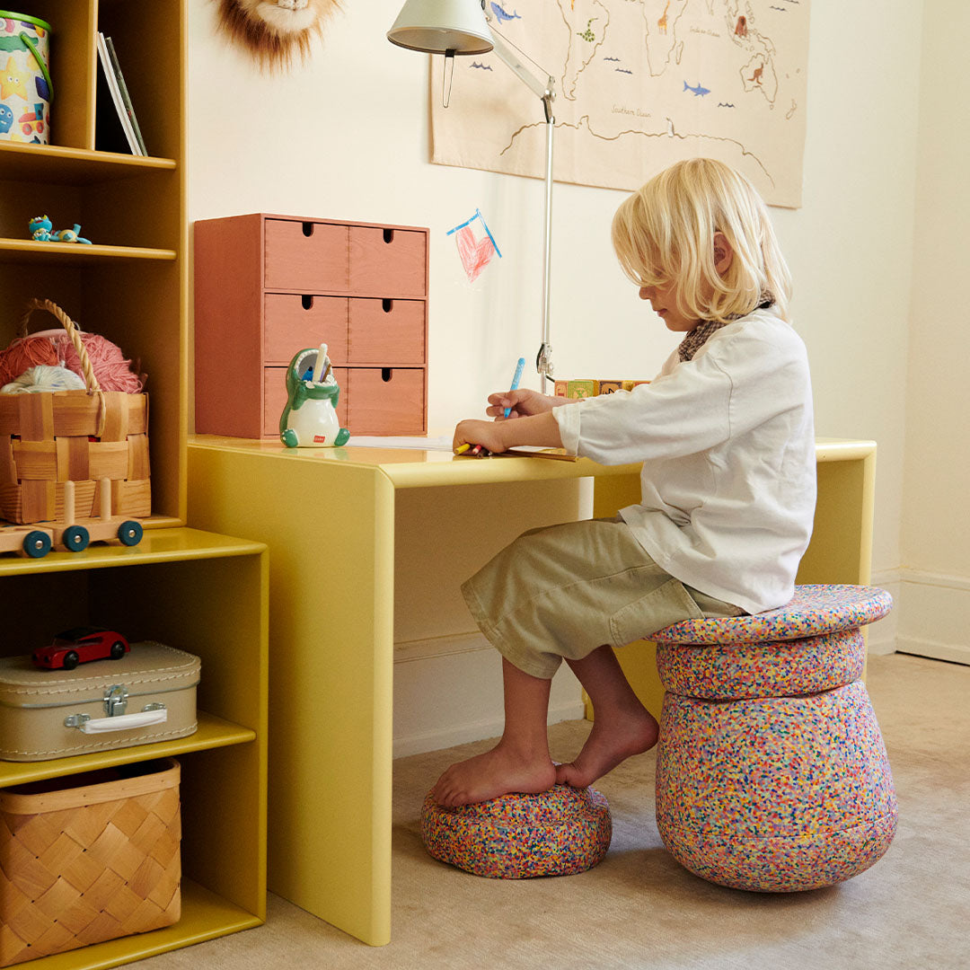 Een kind zit aan een geel bureau op een stapelstein dynamic confetti kruk, met een kleinere stapelstein als voetenbankje.