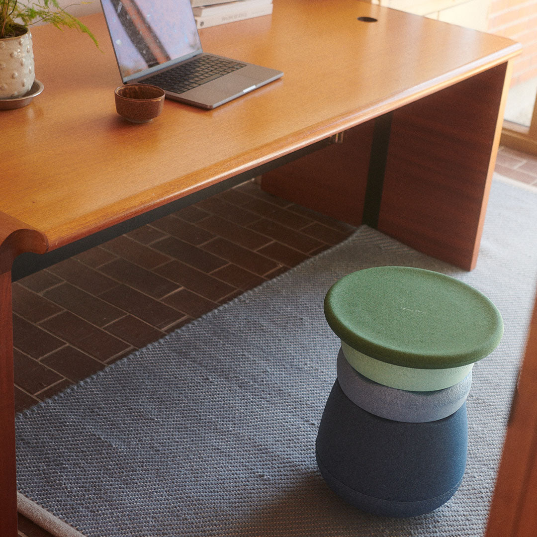 De gestapelde Stapelstein stapelbekers in groen- en blauwtinten staan op een grijs vloerkleed naast een houten bureau.