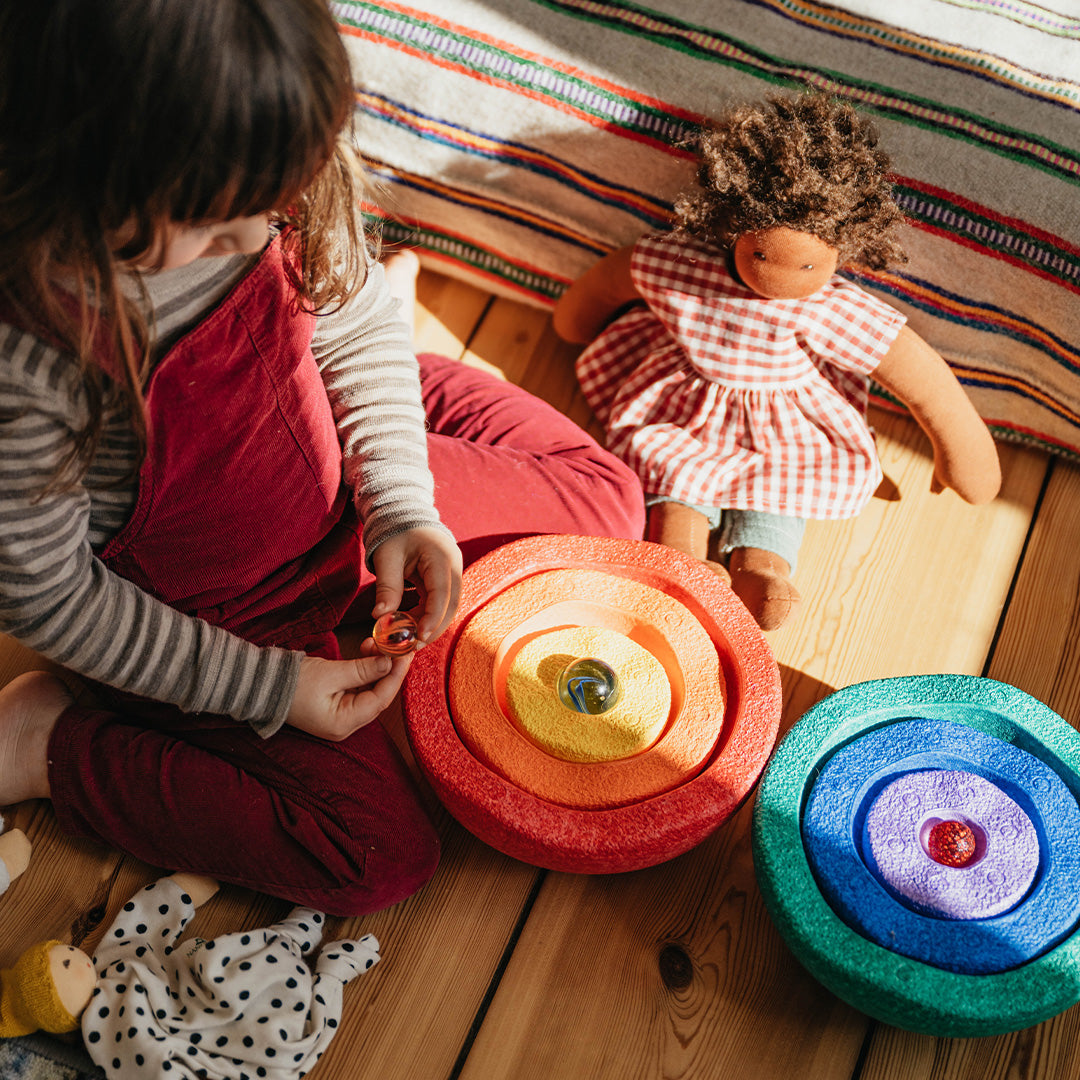 Een kind speelt op een houten vloer met twee sets stapelstein Inside stapelkommen in regenboogkleuren en een knuffelpop.