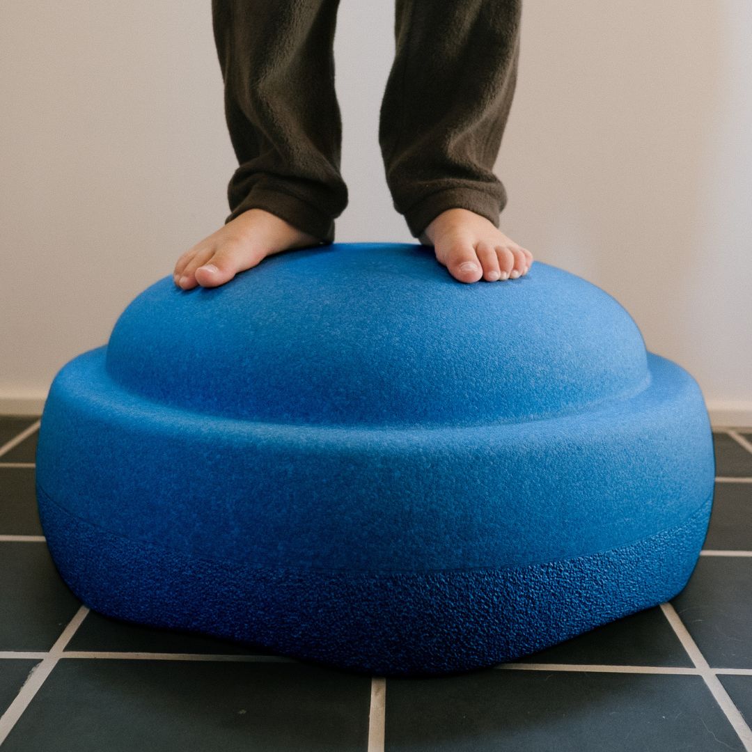Blote kindervoeten staan op een blauw stapelstein schuimrubberblok op een tegelvloer.