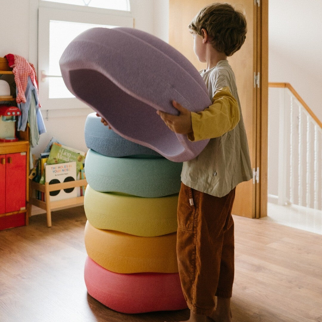 Een kind tilt een paarse stapelstein schuimrubberblok op om deze op een stapel van vijf andere kleurrijke blokken te plaatsen.