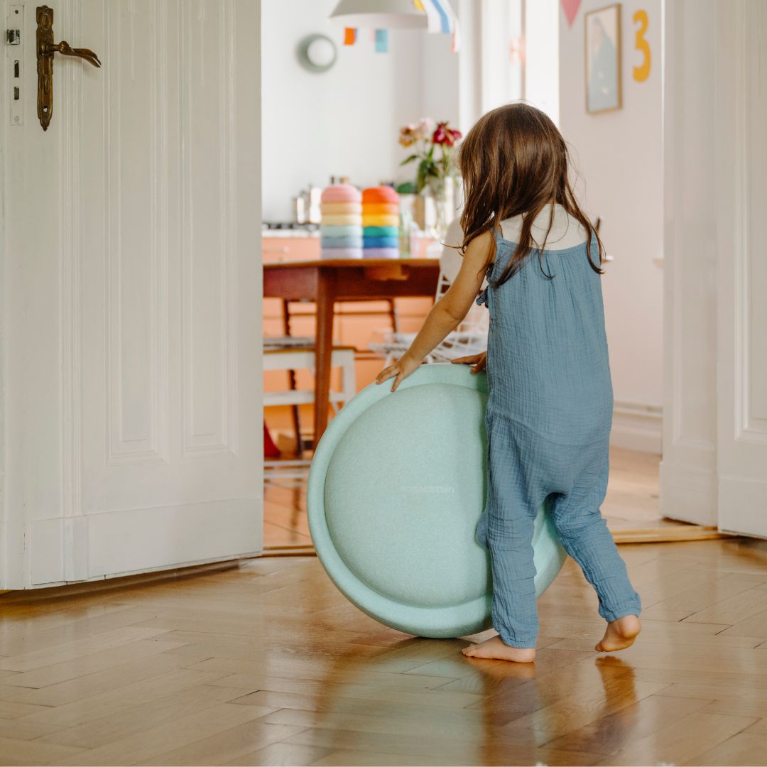 Een kind in een blauwe jumpsuit duwt een lichtgroene stapelstein GROW over een houten vloer.