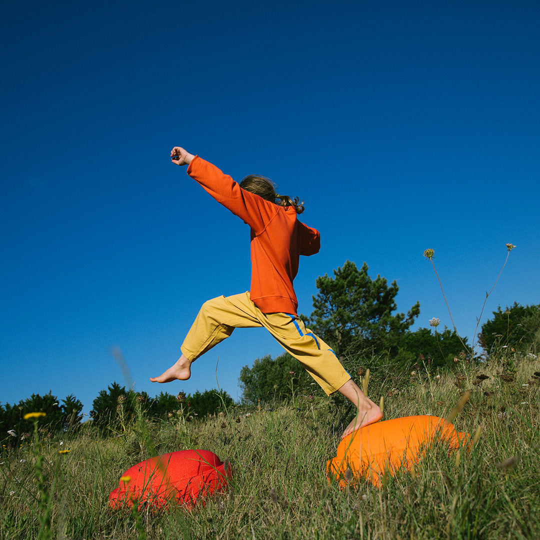 Een kind springt over twee stapelstein schuimrubberblokken, een rode en een oranje, in een grasveld.