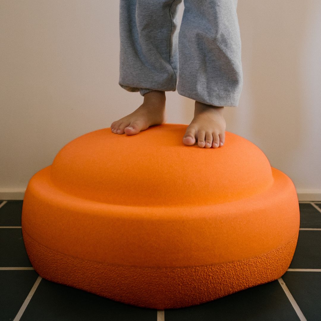 Kindervoeten staan blootsvoets op een oranje stapelstein original GROW schuimrubberblok op een tegelvloer.