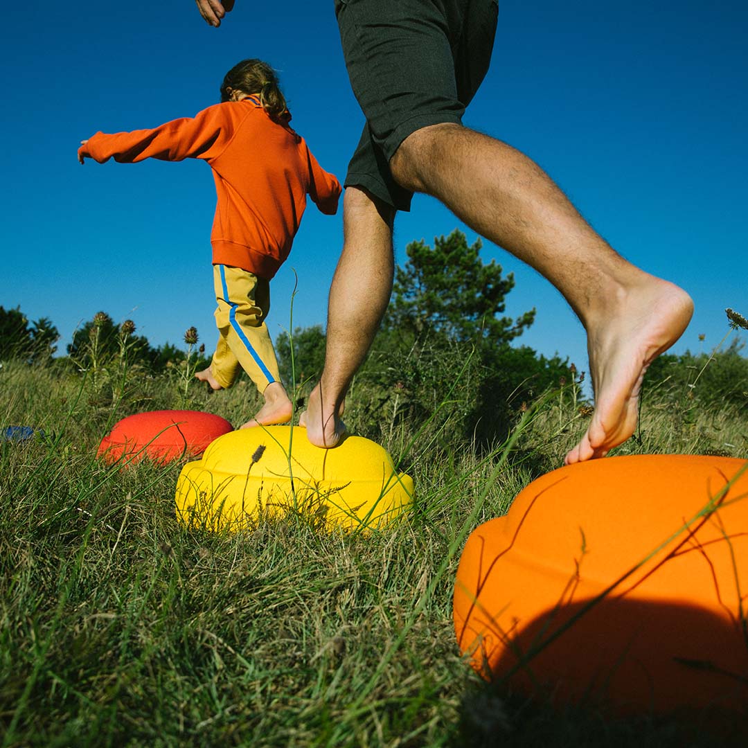 Een kind en volwassene lopen blootsvoets over kleurrijke stapelstein schuimrubberblokken in het gras.