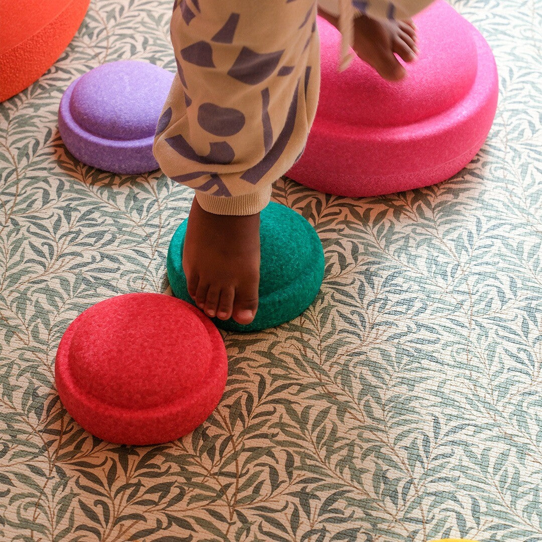 Een kind stapt met blote voet op een groen stapelstein speelelement, met andere elementen op een vloerkleed.