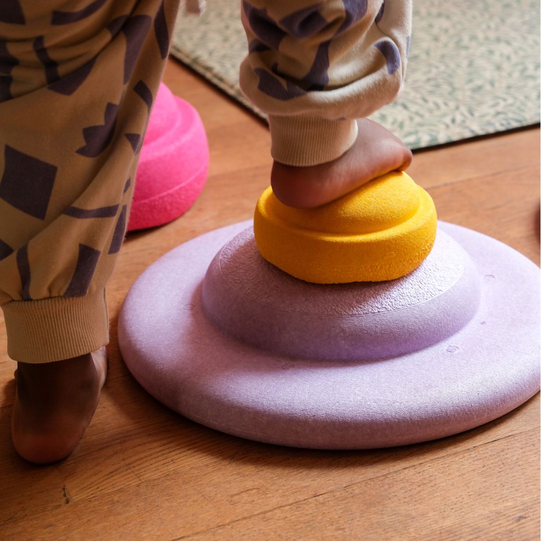 Een kind staat met één voet op een gele stapelstein MINI, die op een paarse stapelsteen ligt.