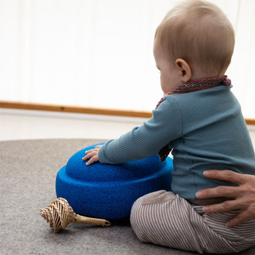 Een baby zit op de grond en raakt een blauw Stapelstein speelelement aan, met een hand die de baby ondersteunt.