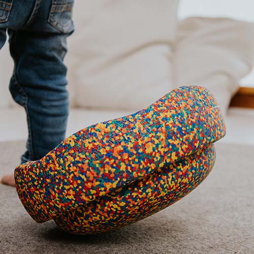 Een kind staat naast een veelkleurig, gespikkeld stapelstein speelelement dat op zijn kant ligt.