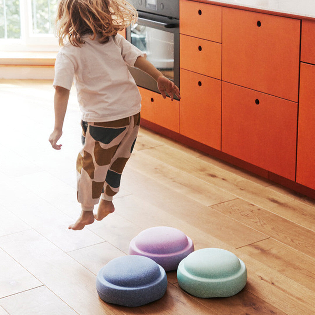 Een kind springt over drie Stapelstein schuimrubberblokken in pastelkleuren op een houten vloer.