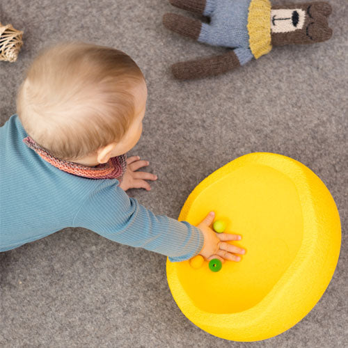 Een baby speelt op de grond met een geel Stapelstein speelelement en kleine gekleurde balletjes.