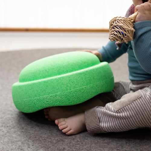 Een baby zit op de grond naast een groen Stapelstein speelelement.