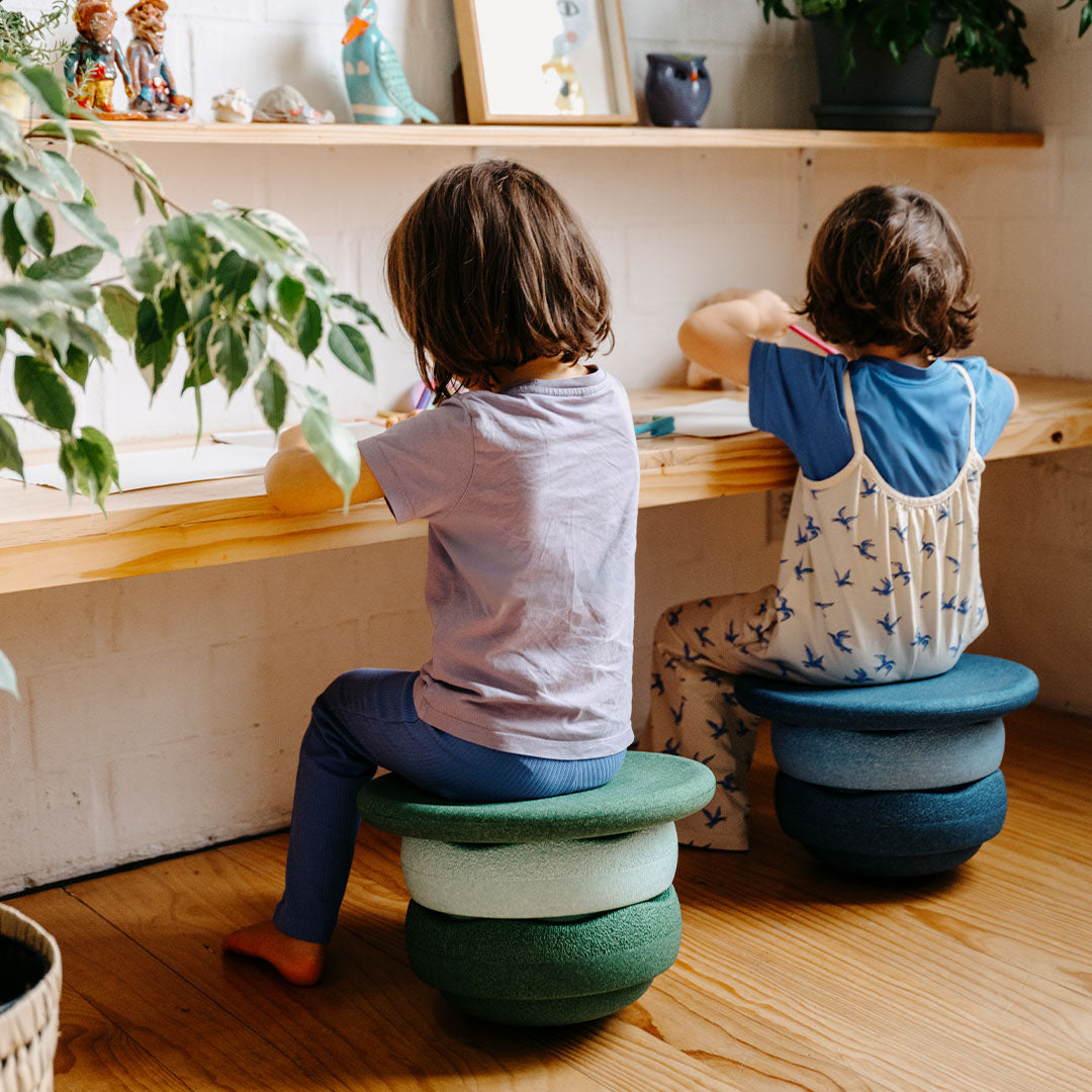 Twee kinderen zitten van achteren gezien op stapels stapelstein balance elementen, één blauw en één groen, aan een houten tafel.
