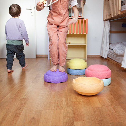 Twee kinderen spelen met stapelstein balansstenen op een houten vloer, één kind staat op een paarse steen.