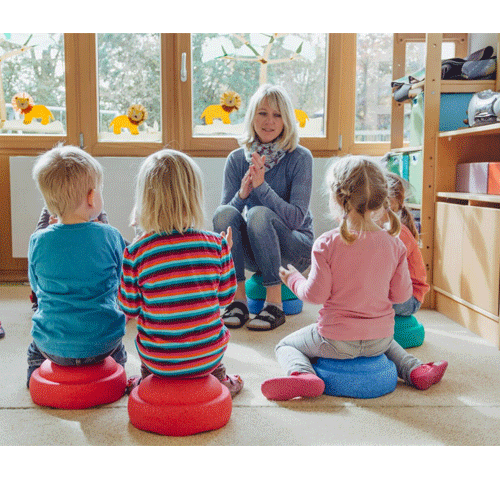 Een groep kinderen zit op kleurrijke Stapelstein speelgoed elementen in een kring met een volwassene.