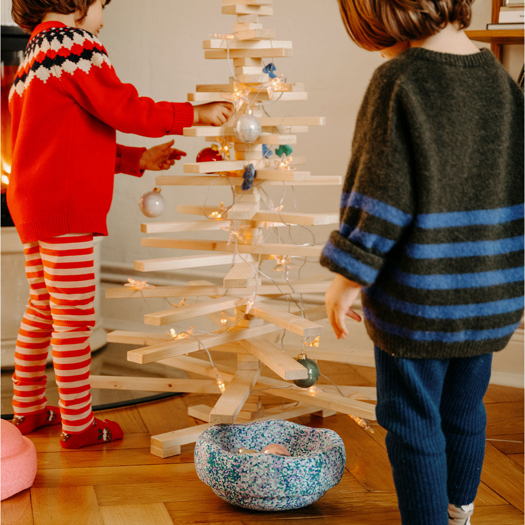 Twee kinderen versieren een houten kerstboom, met een stapelstein element als kom op de voorgrond.