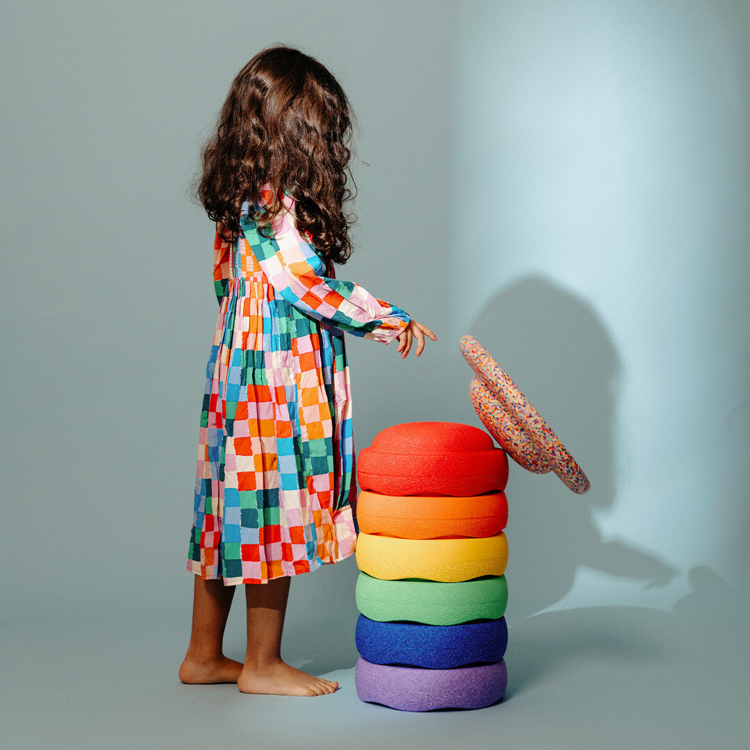 Een kind in een kleurrijke jurk staat naast een stapel stapelstein regenboog balance boards.