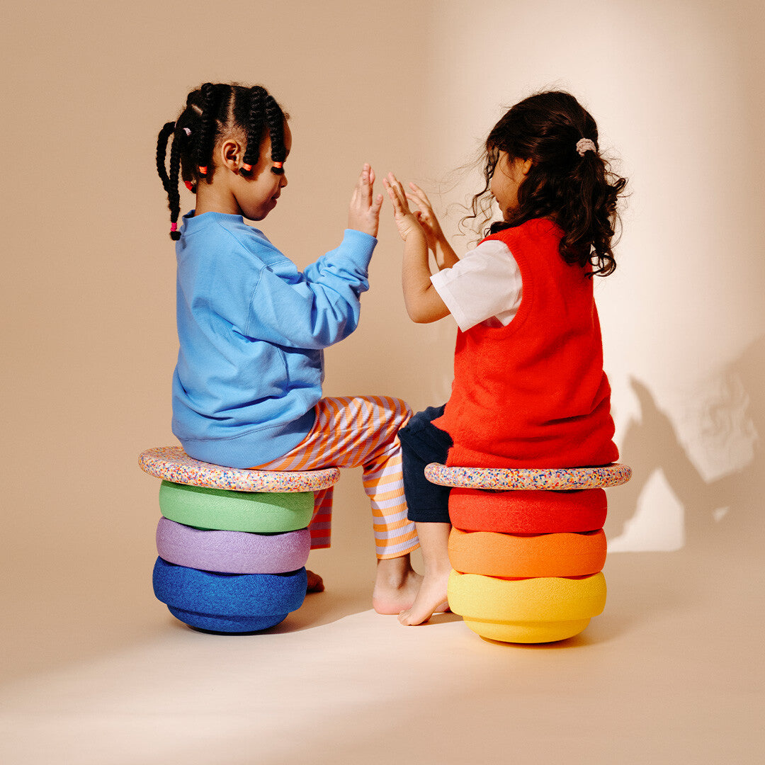 Twee kinderen zitten op stapels stapelstein balance boards en spelen een klapspel.