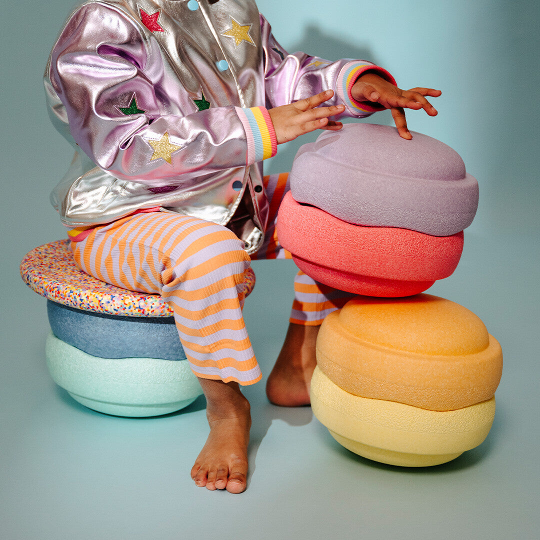 Een kind zit op een stapel Stapelstein balance boards in pastelkleuren, met een tweede stapel ernaast.