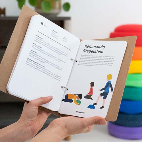 De geopende pagina's van het stapelstein spellenboek tonen spelinstructies met illustraties van kinderen.