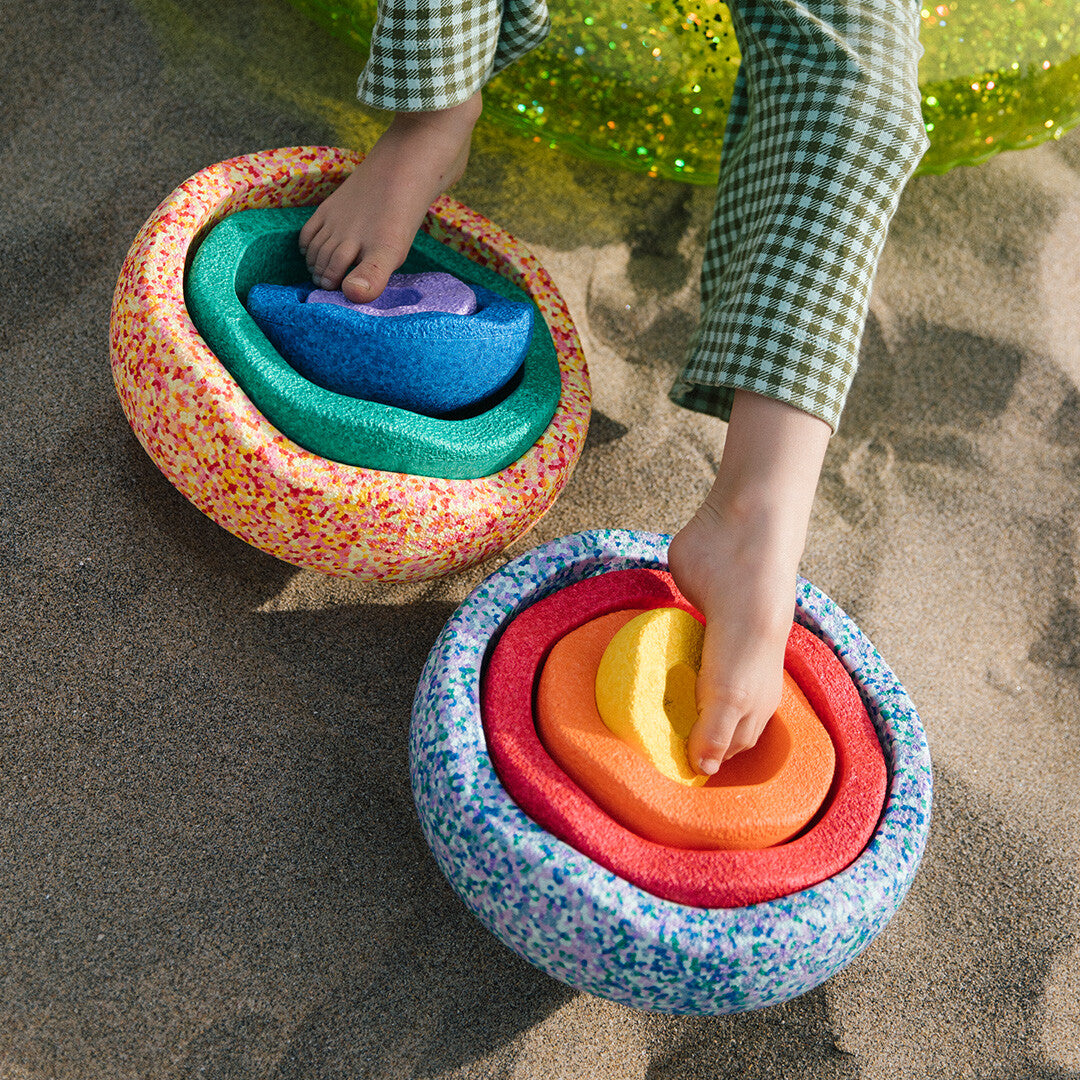 Kindervoeten spelen op het zand met twee geneste stapelstein summer rainbow sets, waarbij een geel element wordt geplaatst.