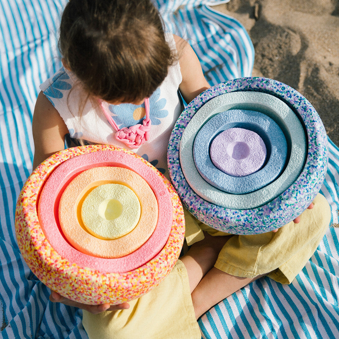 Een kind zit op het strand en houdt twee sets stapelspeelgoed in pastelkleuren vast.