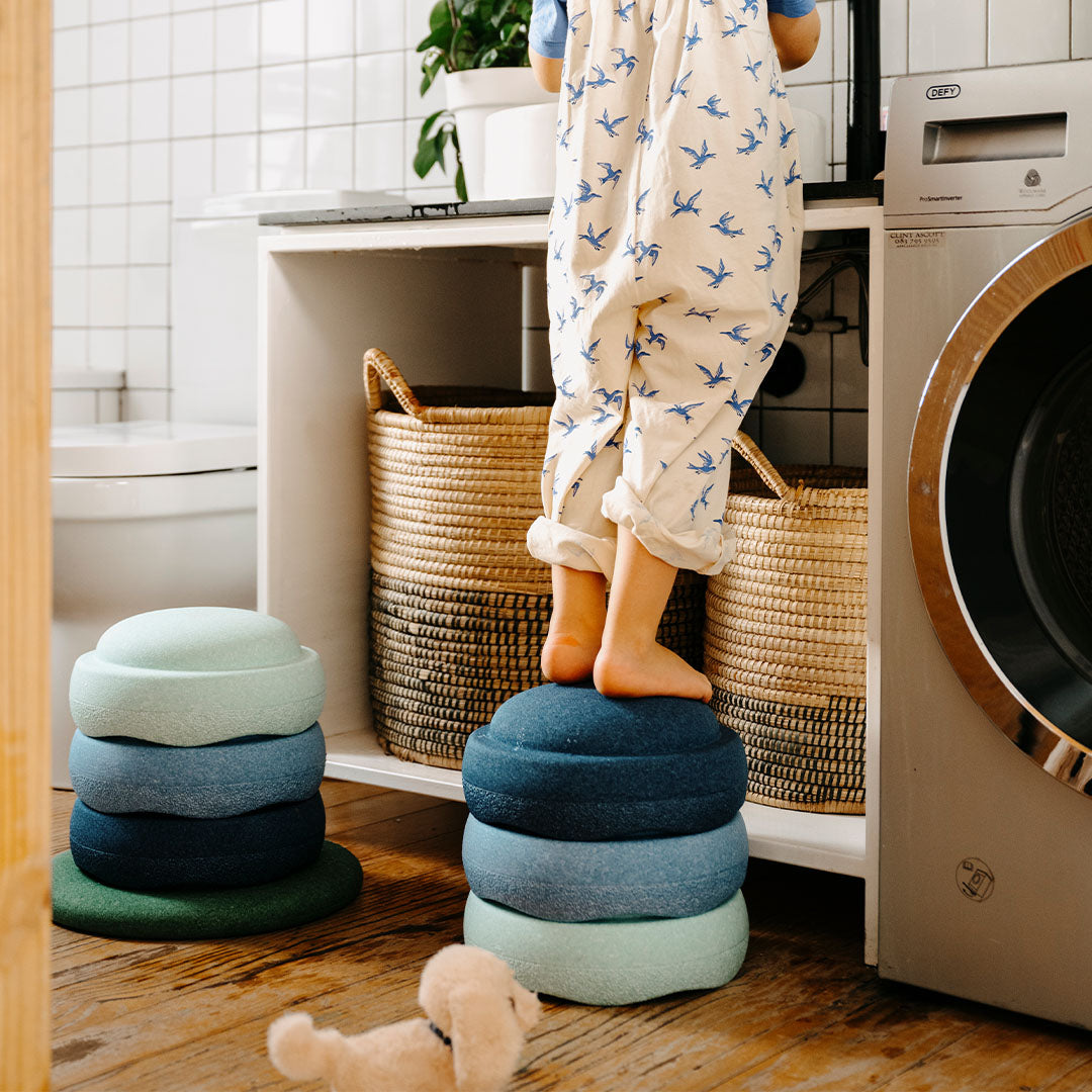 Een kind staat op een stapel stapelstein elementen in blauw- en groentinten in een badkamer.
