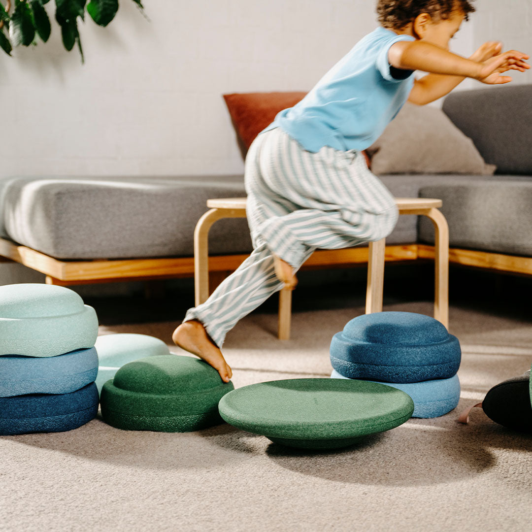 Een kind springt over een tafeltje, met stapelstein balanceerelementen in groene en blauwe tinten op de vloer.