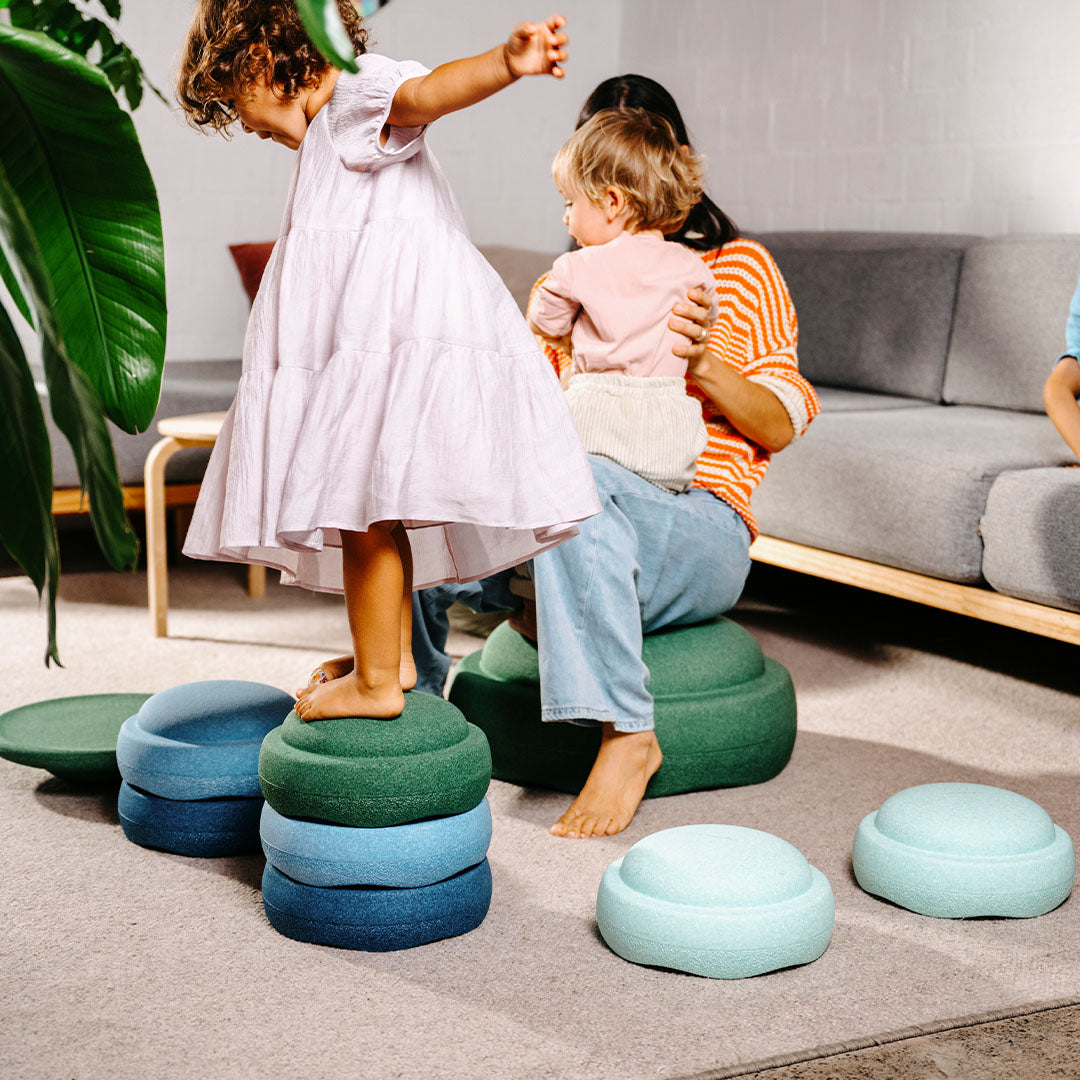Twee kinderen en een volwassene spelen binnenshuis met stapelstein balanceerelementen in groene en blauwe kleuren.