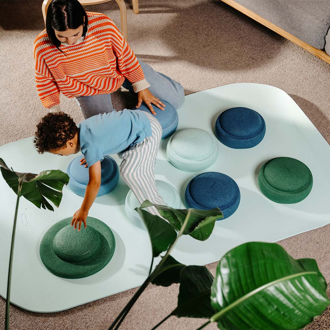 Bovenaanzicht van een kind en een volwassene die spelen met stapelstein balanceerelementen in groene en blauwe tinten op een lichtblauwe mat.