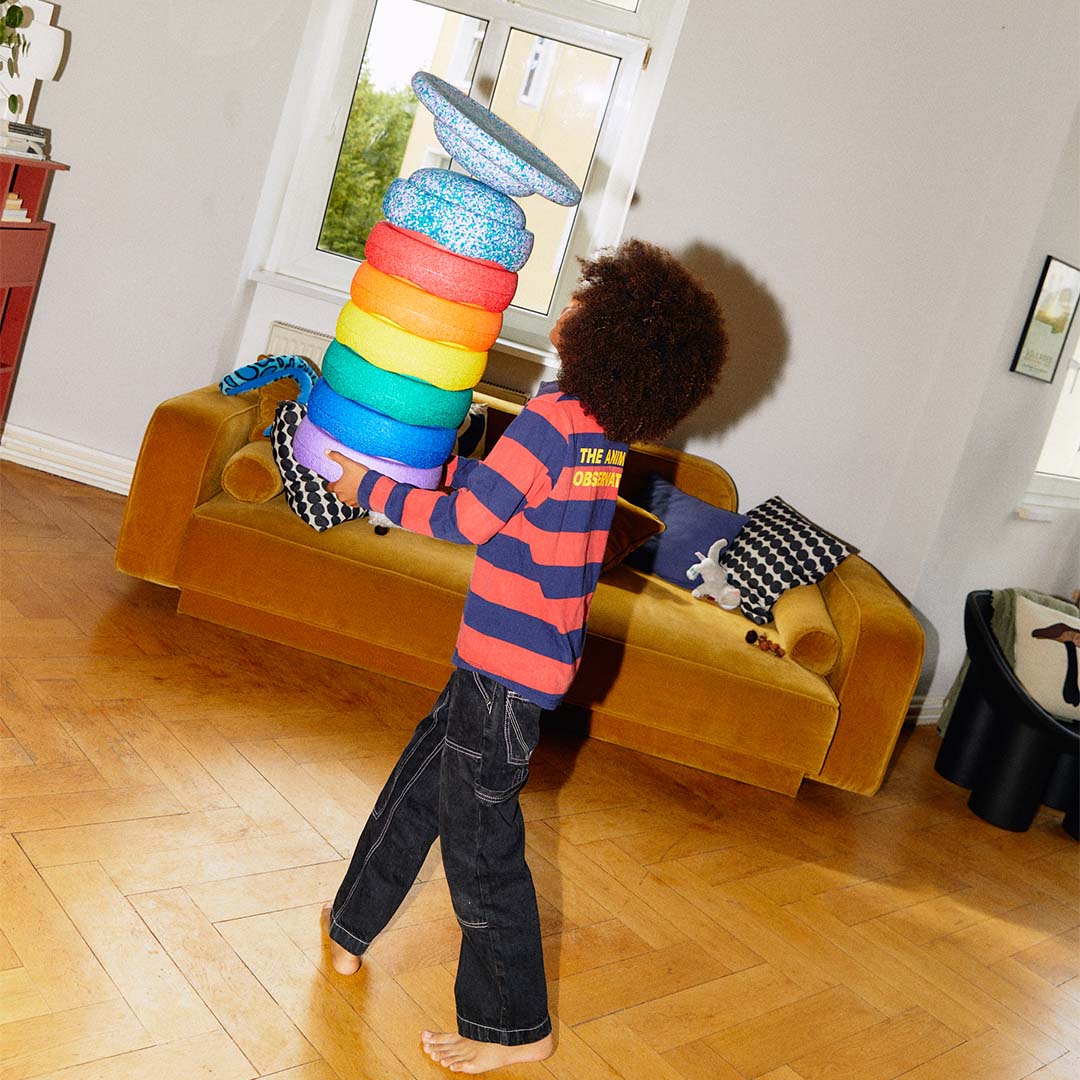 Een kind draagt een hoge stapel regenboogkleurige stapelstein schuimrubberblokken door een kamer.