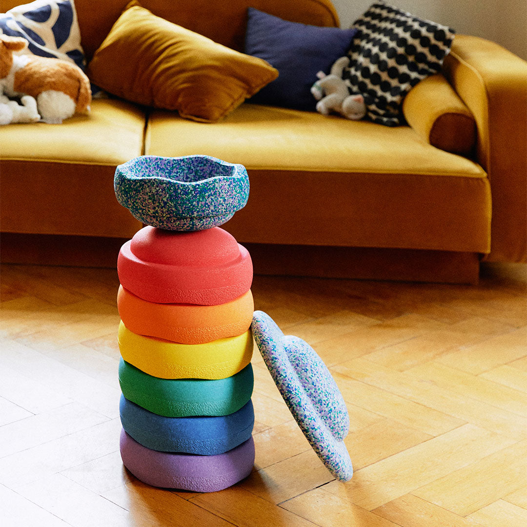 Een stapel regenboogkleurige stapelstein schuimrubberblokken met een gespikkeld blauw element erbovenop en ernaast, in een woonkamer.