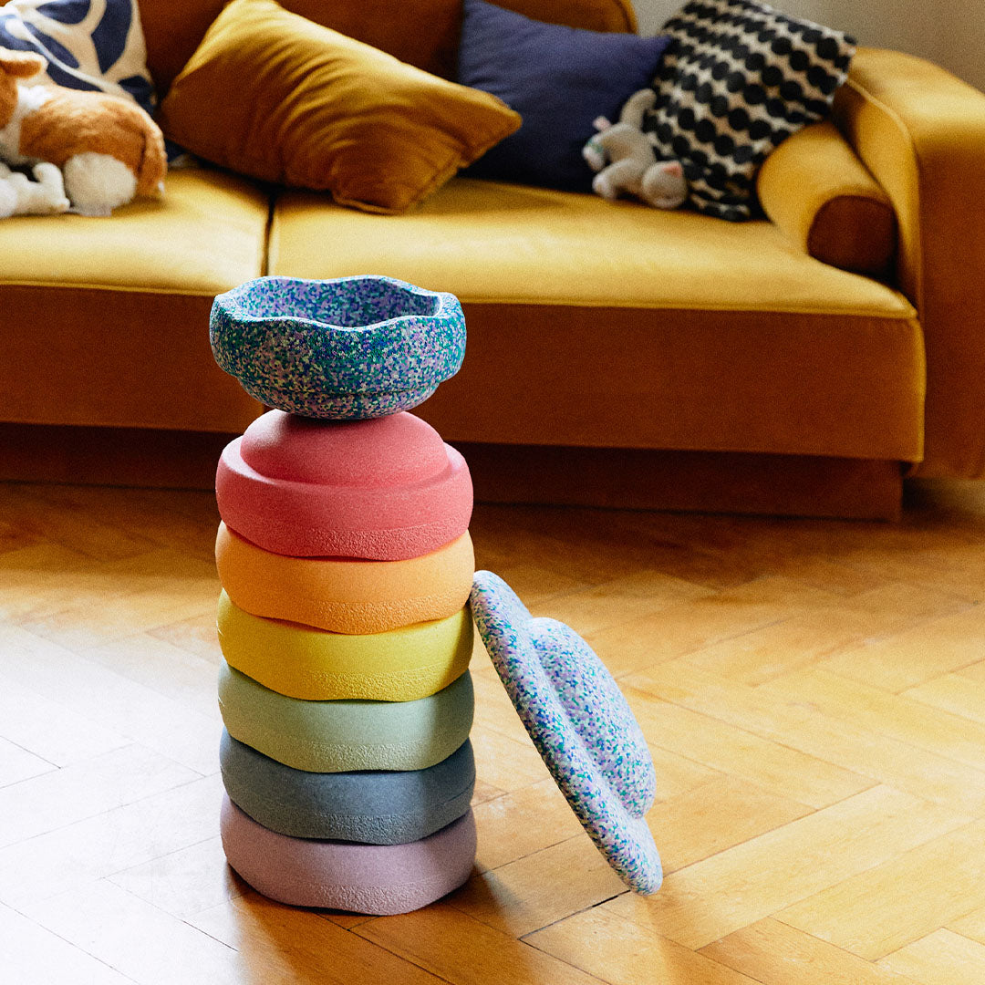 Een stapel stapelstein schuimrubberblokken in pastel regenboogkleuren staat op een houten vloer voor een gele bank.