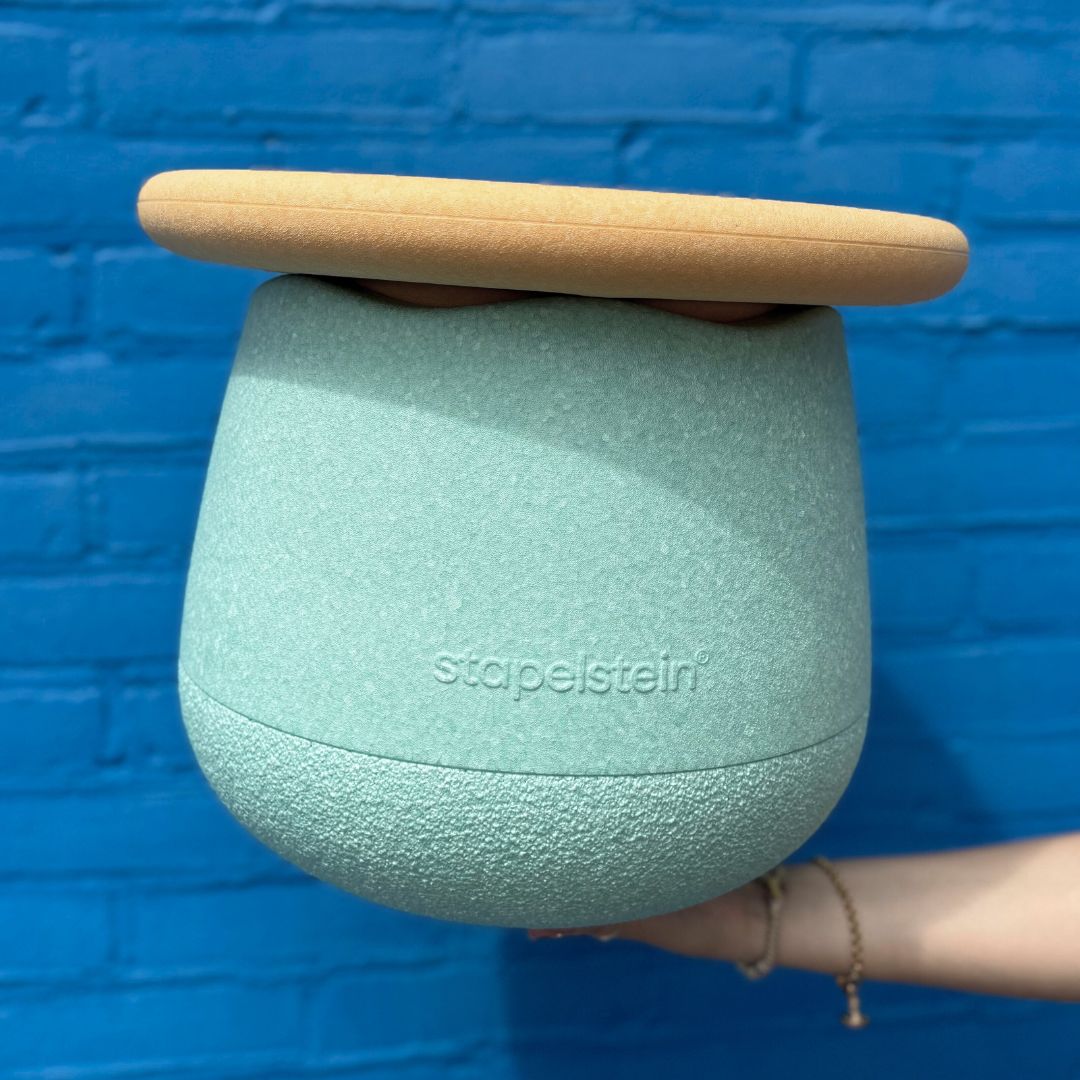 Een hand houdt het Stapelstein dynamic small fitnessspeeltje met lichtblauwe basis en oranje bovenkant vast voor een blauwe muur.