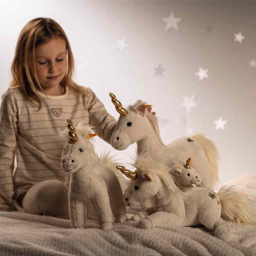 Een meisje zit op bed en kijkt naar drie Steiff pluchen eenhoorns van verschillende formaten.